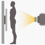 Silhouetted person standing by a wall-mounted imaging device emitting a yellow beam toward their torso, illustrating a scan.