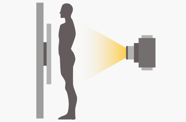 Silhouetted person standing by a wall-mounted imaging device emitting a yellow beam toward their torso, illustrating a scan.