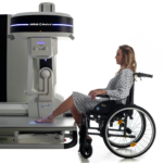 Woman in a wheelchair sits beside a large medical imaging scanner, preparing to undergo an exam; the machine has a dark band and blue lights and is labeled aptitude.