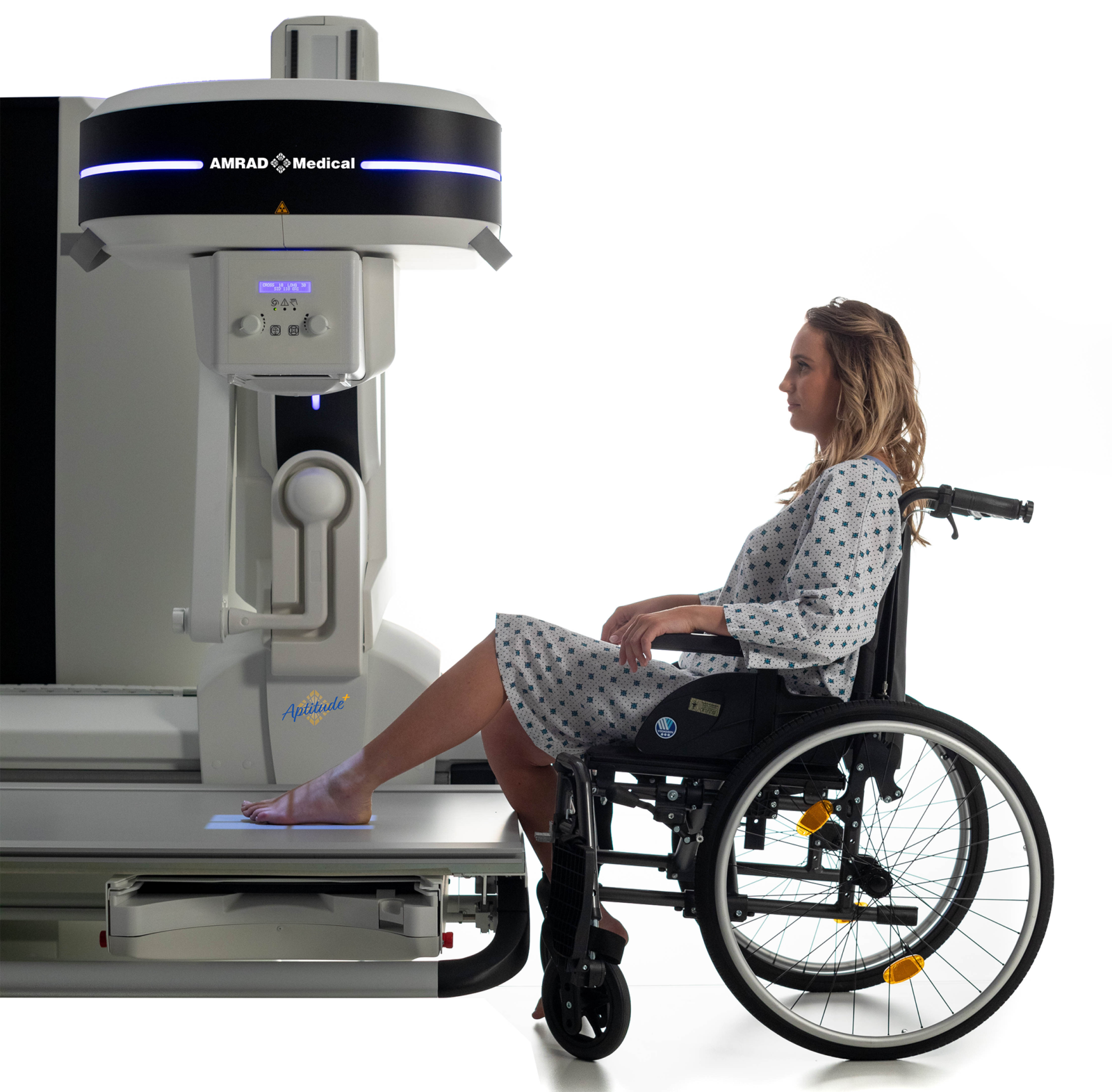 Woman in a wheelchair sits beside a large medical imaging scanner, preparing to undergo an exam; the machine has a dark band and blue lights and is labeled aptitude.