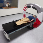 Patient lying on a ct scanner table inside a large circular gantry, preparing for imaging. Variance: radiology tech monitors from an adjacent room through a window.