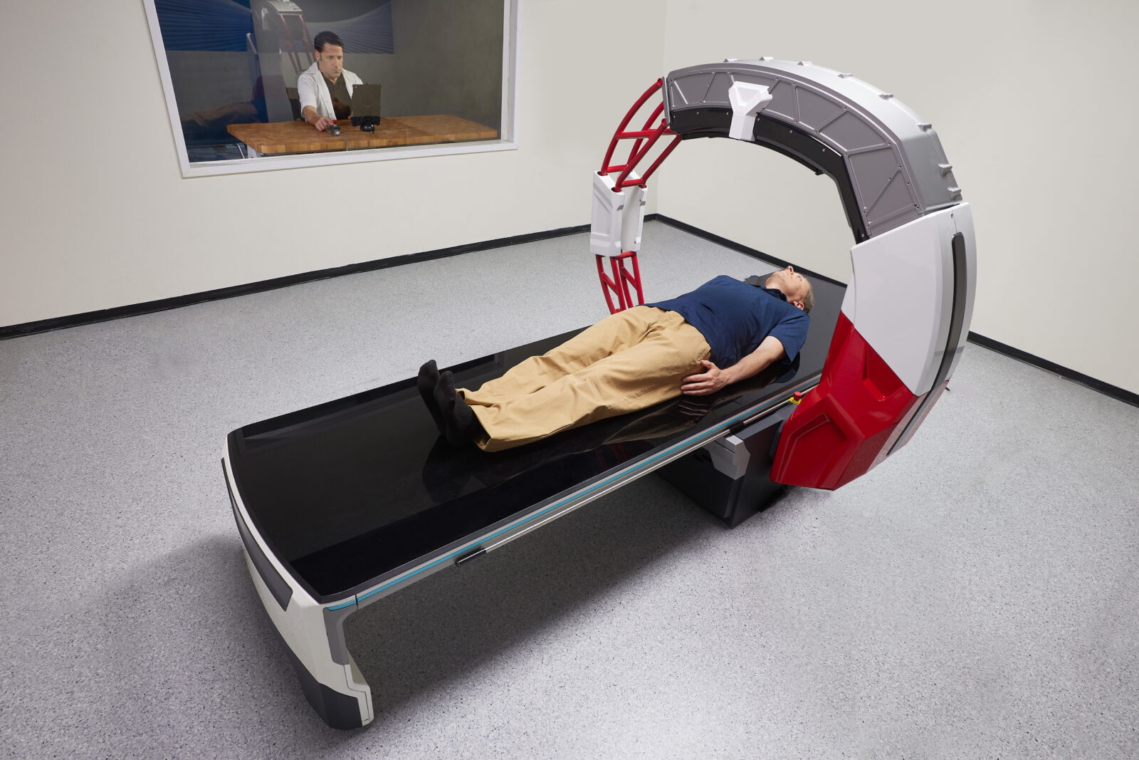 Patient lying on a ct scanner table inside a large circular gantry, preparing for imaging. Variance: radiology tech monitors from an adjacent room through a window.
