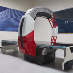 Medical imaging scanner with a circular gantry and red support frame in a showroom, nanox logo visible on a blue wall behind it. “