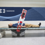 Man lying on a medical imaging table with a red equipment frame tilted over him against a blue nanox branded wall backdrop, in a clinical setting.