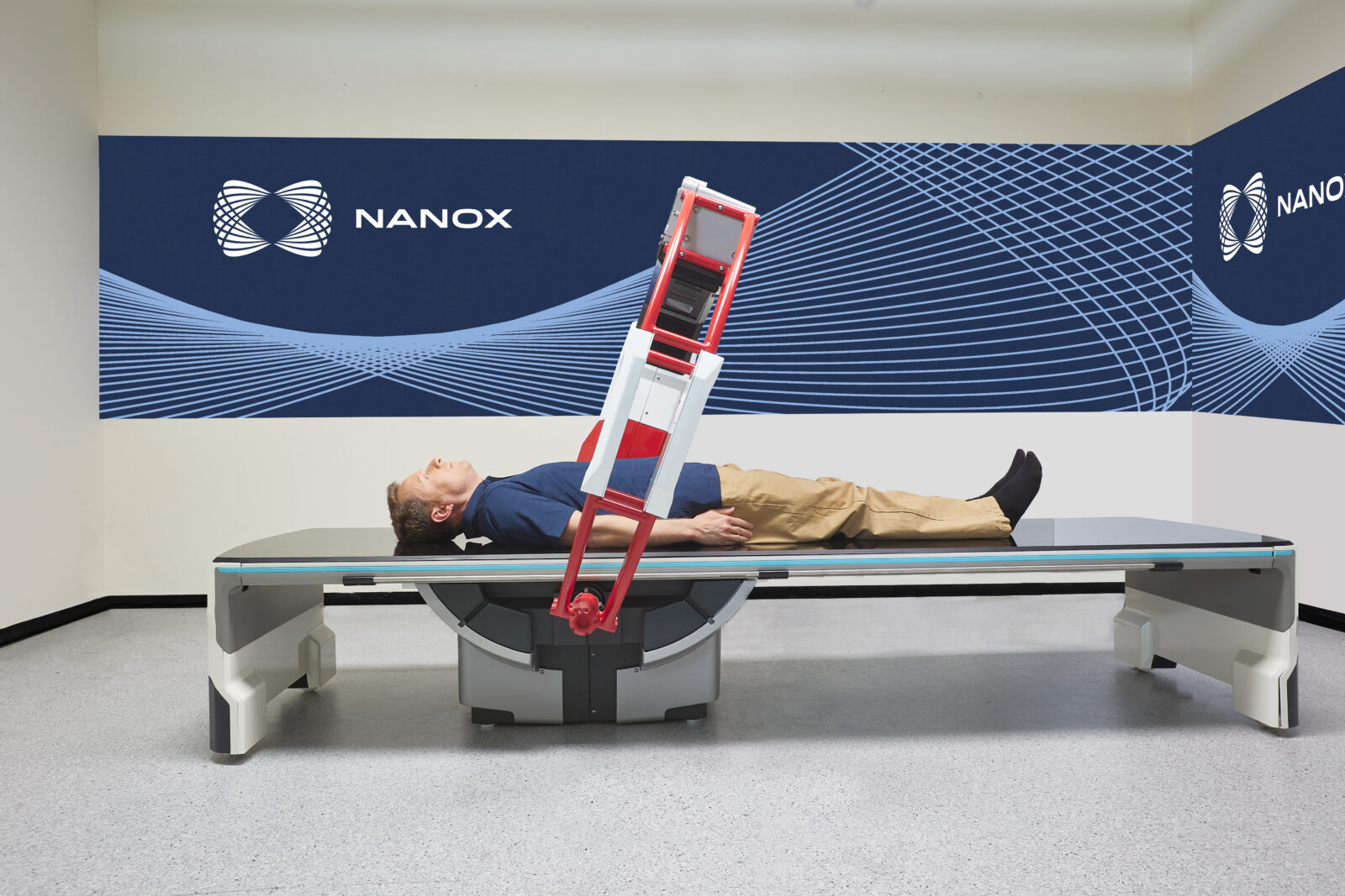 Man lying on a medical imaging table with a red equipment frame tilted over him against a blue nanox branded wall backdrop, in a clinical setting.