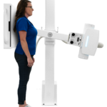 Woman in blue shirt stands beside a tall white medical imaging machine with an adjustable arm/gantry.