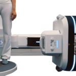 Medical linear accelerator in a radiotherapy setup, with a patient standing on the treatment platform beside the machine labeled amrad medical.