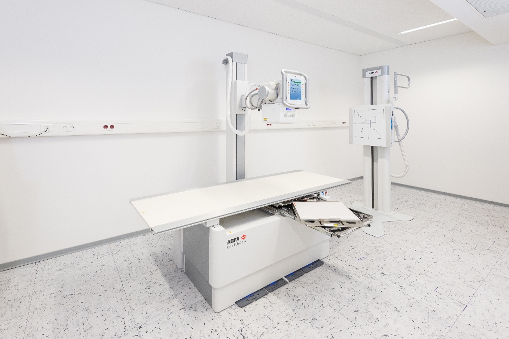 A hospital radiography room with an adjustable x-ray table and wall-mounted equipment, including a monitor and imaging device on a stand behind the table, all in white.