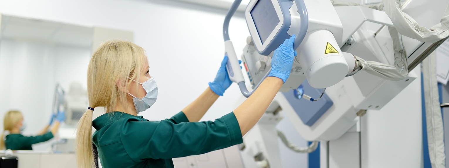 Imit-home-xray - Imperial Imaging Technology Clinician in a mask and gloves adjusts a large medical imaging machine in a bright hospital room, touching its controls.