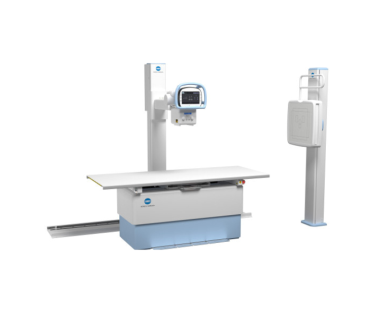 Medical imaging setup featuring an x-ray table with a vertical upright scanner and a separate control console on a tall column to the left; white and light-blue color scheme.