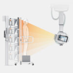 Patient stands at a vertical imaging stand while a robotic c-arm x-ray device emits a cone of radiation toward the patient.
