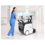 Nurse in blue scrubs pushes a medical imaging robot cart through a bright hospital corridor.