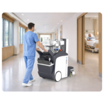 Healthcare worker in blue scrubs operates a wheeled portable medical device in a bright hospital hallway toward patient rooms. 2024 model included. (informative) - concise alt text for screen readers.