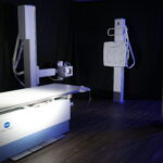 Medical imaging room featuring a konica minolta radiography table and vertical x-ray unit against dark backdrop with blue lighting.