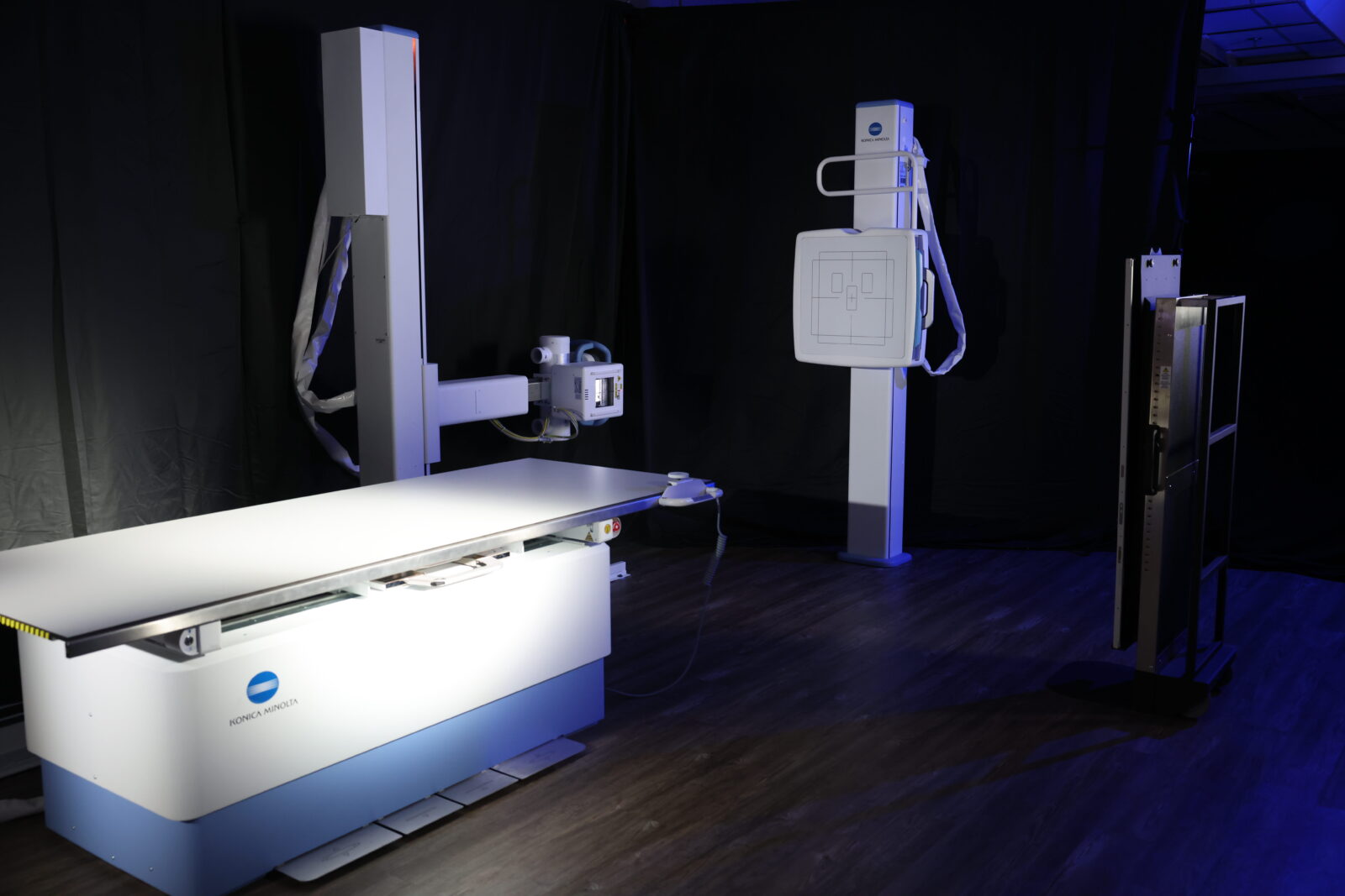 Medical imaging room featuring a konica minolta radiography table and vertical x-ray unit against dark backdrop with blue lighting.