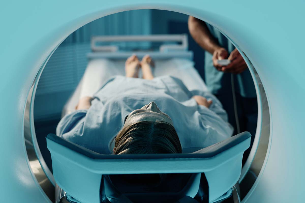 Radiographer-working-with-female-patient A person is lying on a hospital bed, entering an MRI scanner. They are covered with a blanket, and a medical professional is adjusting controls nearby. The room has a clinical, sterile environment, and the scanner's circular opening is prominent.