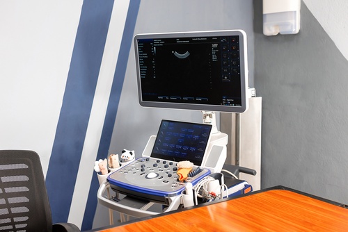Portable ultrasound machine with monitor and control panel on a wheeled cart beside a desk in a clinic room.