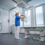 Radiology technician in blue scrubs adjusts controls on an overhead x-ray machine beside an examination table in a bright imaging room.