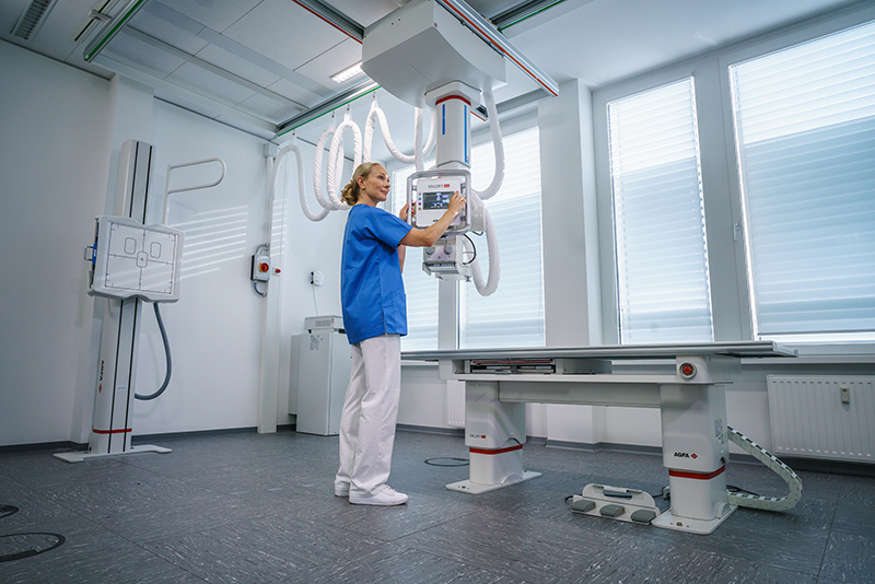 Radiology technician in blue scrubs adjusts controls on an overhead x-ray machine beside an examination table in a bright imaging room.