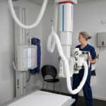 Radiologic technologist beside a ceiling-mounted x-ray gantry, preparing to image a patient on the examination table in a clinical imaging room