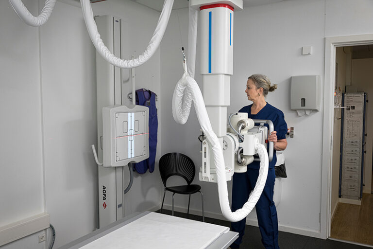 Radiologic technologist beside a ceiling-mounted x-ray gantry, preparing to image a patient on the examination table in a clinical imaging room