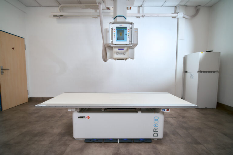 Ceiling-mounted radiography machine above a long x-ray table in a clinical room, with agfa branding on the base table