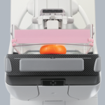 Close-up of a medical imaging device's loading tray, featuring a pink insert and a transparent bin holding an orange ball.