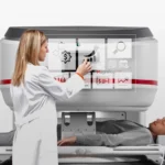 Clinician operates a large medical imaging/treatment machine over a patient lying on a table for radiotherapy or scanning.