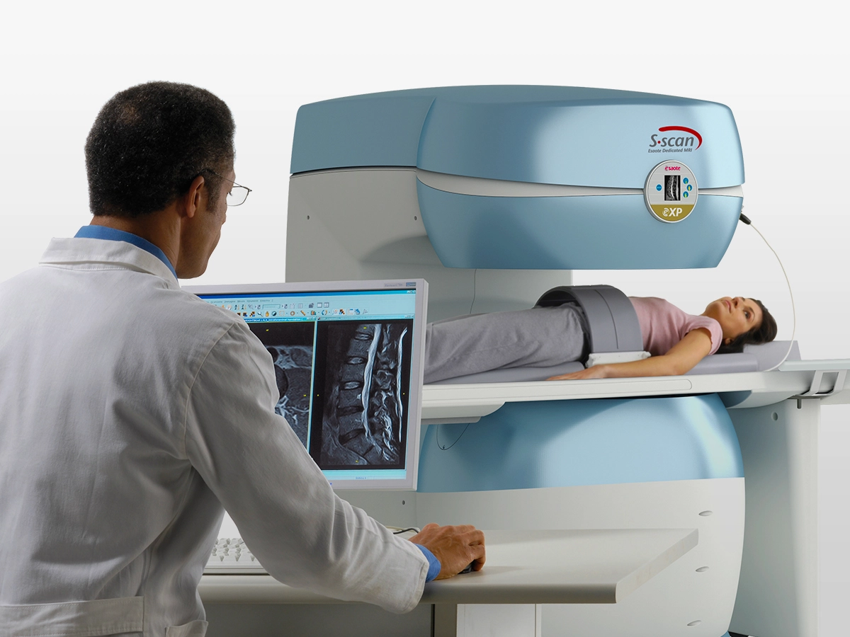 Technician in a white coat operating an mri machine while a patient lies on the scanner table awaiting imaging. A computer monitor shows spinal mri scans nearby.