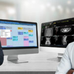 Two healthcare professionals stand with smiling faces in front of dual computer displays showing a clinic scheduling calendar and medical imaging tools.